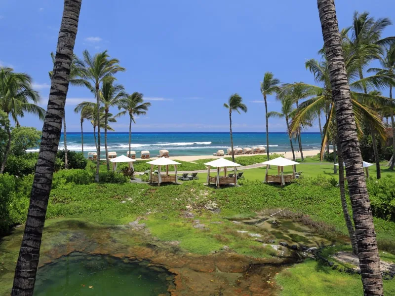 A Kohala Coast resort lawn framed by coconut palms, white shade cabanas, an ancient fishpond, and turquoise ocean beyond.