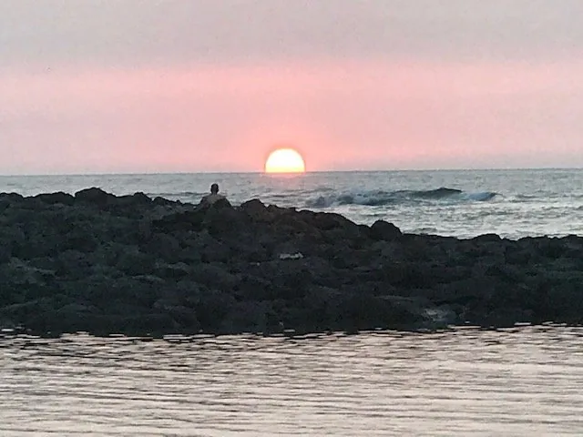 A Kona sunset dropping onto the ocean horizon, one person silhouetted on the lava rocks in the foreground.